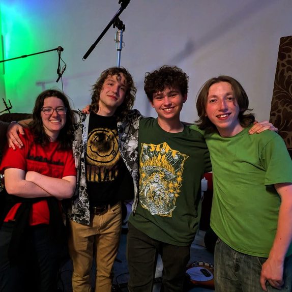 The four members of Across 35th, freshmen at West Seattle High School, stand shoulder to shoulder with arms around each other, laughing in their rehearsal space under green and blue stage lighting.