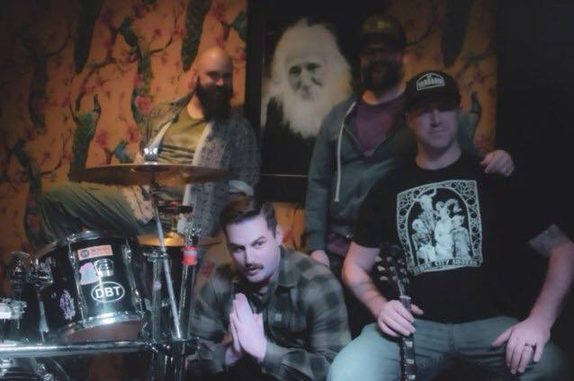 Cabeza gathered around a drum kit at a bar venue in Boise, dark floral wallpaper behind them — one member crouched by the kit, one kneeling with hands clasped, and two standing, one with his arm around the other's shoulder.