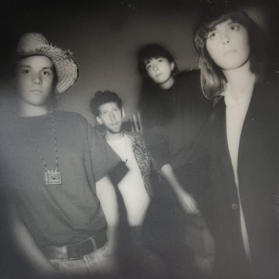 Black-and-white portrait of Capping Day's four members standing close together against a dark backdrop — one in a straw hat with a beaded chain necklace, one in an open shirt, and two women in dark blazers, one with a bob and one with long wavy hair.