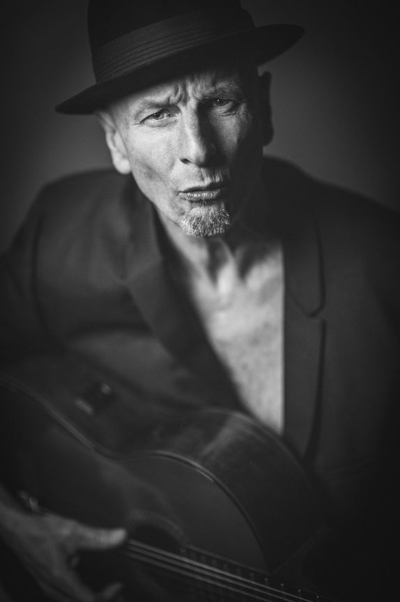Black-and-white studio portrait of Gary Minkler wearing a fedora, holding an acoustic guitar.