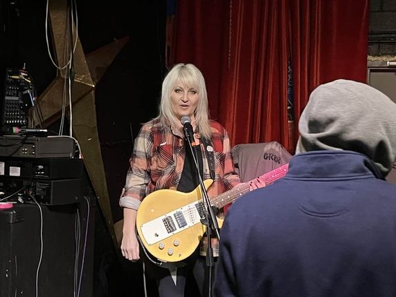 Lisa Koenig singing and playing a mustard-yellow Les Paul guitar into a mic stand, red stage curtain behind her.
