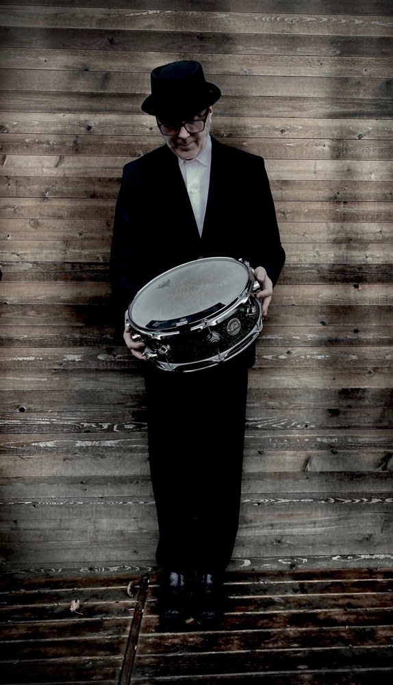Jeff Parkhurst standing in a top hat and dark suit, holding a snare drum, against a weathered wooden wall.