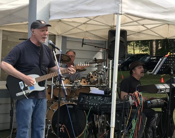 Joe Cason on guitar and vocals performing live outdoors under an event tent, with a drummer and keyboardist behind him.