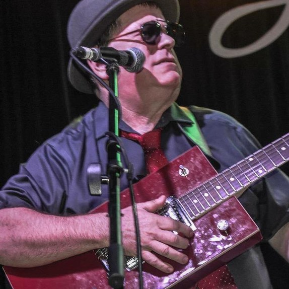Joey Kline in a flat cap and sunglasses, singing into a microphone while playing a red cigar box slide guitar on stage