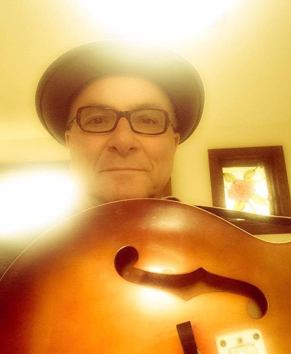 Kaz Murphy in a fedora and glasses, taking a backlit selfie while holding a sunburst archtop guitar