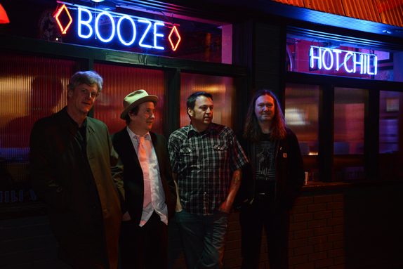 The four members of King County Queens standing outside a bar at night, under neon BOOZE and HOT CHILI signs