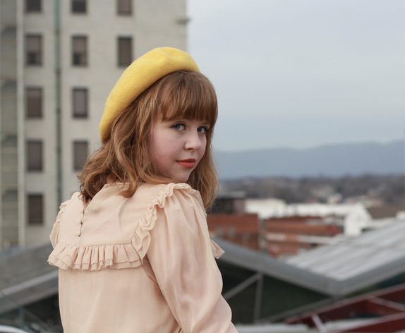Olivia Bloch in a yellow beret and pale pink ruffled blouse, looking back over her shoulder on a rooftop, with an apartment block and distant hills behind her
