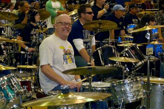 Peter Barnes smiling behind his drum kit, surrounded by other drummers at a group drumming event.