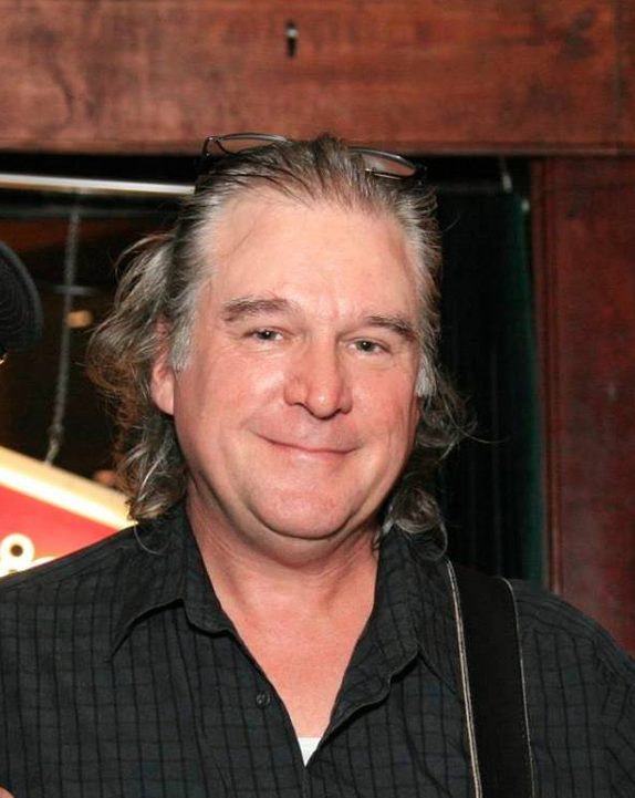 Rich Hinklin, smiling, with glasses pushed up on his head and a guitar strap over his shoulder