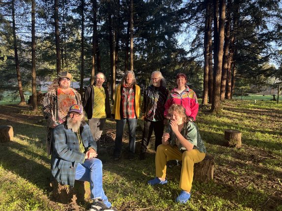 The Hinges posing outdoors among evergreen trees, five members standing and two seated on tree stumps in front.