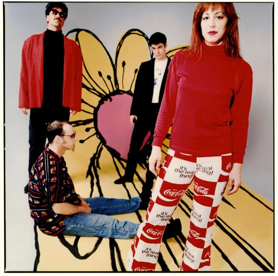 The Holidays in 1992 — four band members in front of a painted flower backdrop, the singer in a red top and Coca-Cola checkerboard trousers