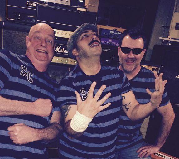 The Shaken Growlers — Mike Dippery, Phil Bentz, and Jeff Hood posing together in matching striped SG shirts backstage amid a wall of guitar amps.