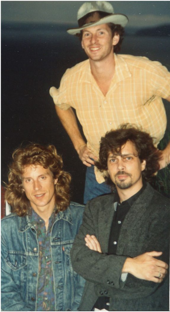Three members of Village Green outdoors at dusk near water — two seated in front, one in a denim jacket and one in a dark blazer with arms crossed, and one standing behind them in a straw hat and yellow plaid shirt