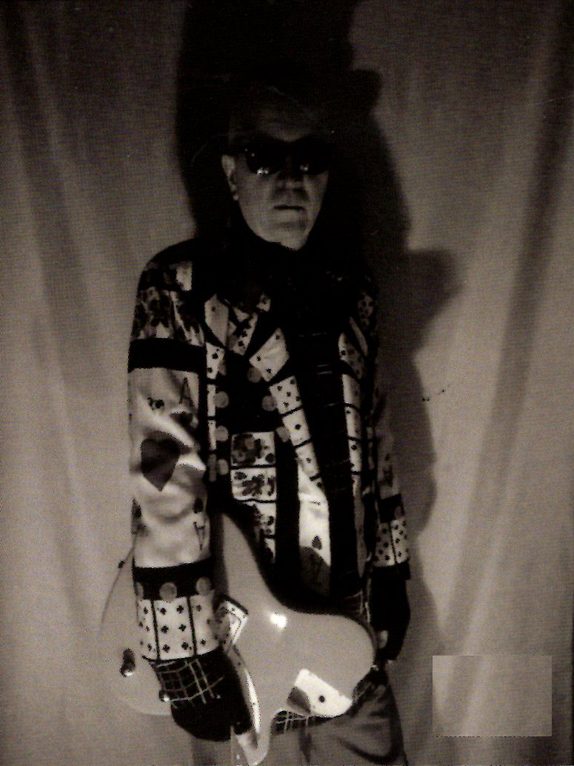 Grady Eddins in dark sunglasses and a playing-card-patterned jacket, holding a white electric guitar, posed against a curtain backdrop.