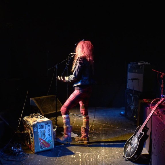 Zelda Starfire performing on stage, guitar in hand and back to the camera, red plaid leggings lit under stage lights