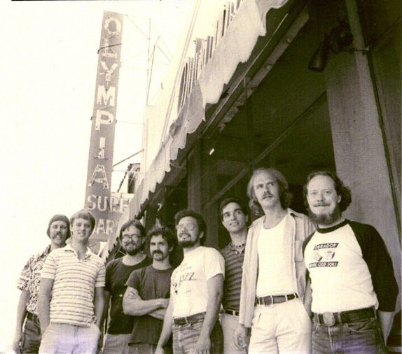 Obrador in San Francisco, 1983 - the seven-piece standing in a row on the sidewalk outside the Olympia Surplus Market, one member in an Obrador shirt.