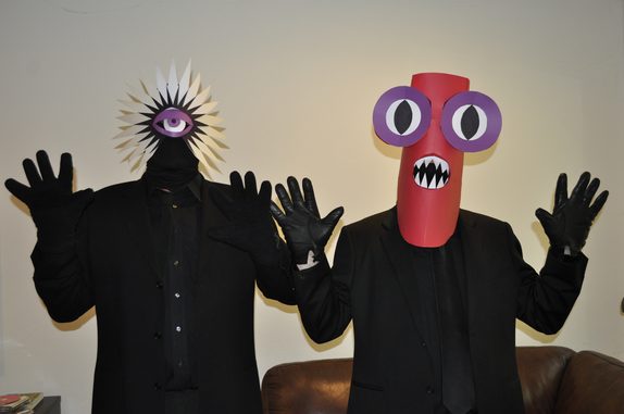 Pop Star Devils — two figures in black suits and gloves with hands raised, one in a white starburst mask with a single eye, the other in a tall red mask with purple goggle eyes and a fanged mouth. Photo by Howie.